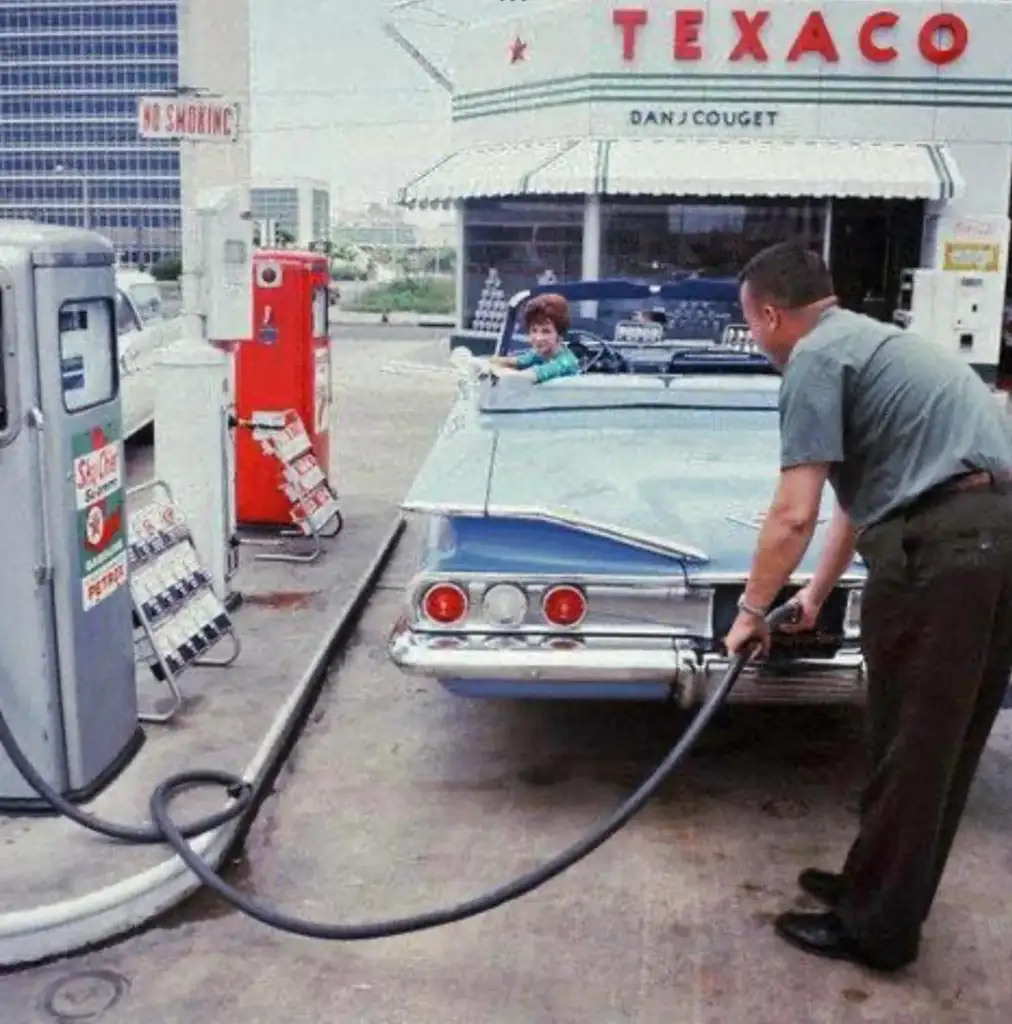 A vintage scene at a Texaco gas station with a man refueling a classic blue car. A person sits inside the convertible. The station features an old-style gas pump and signage.