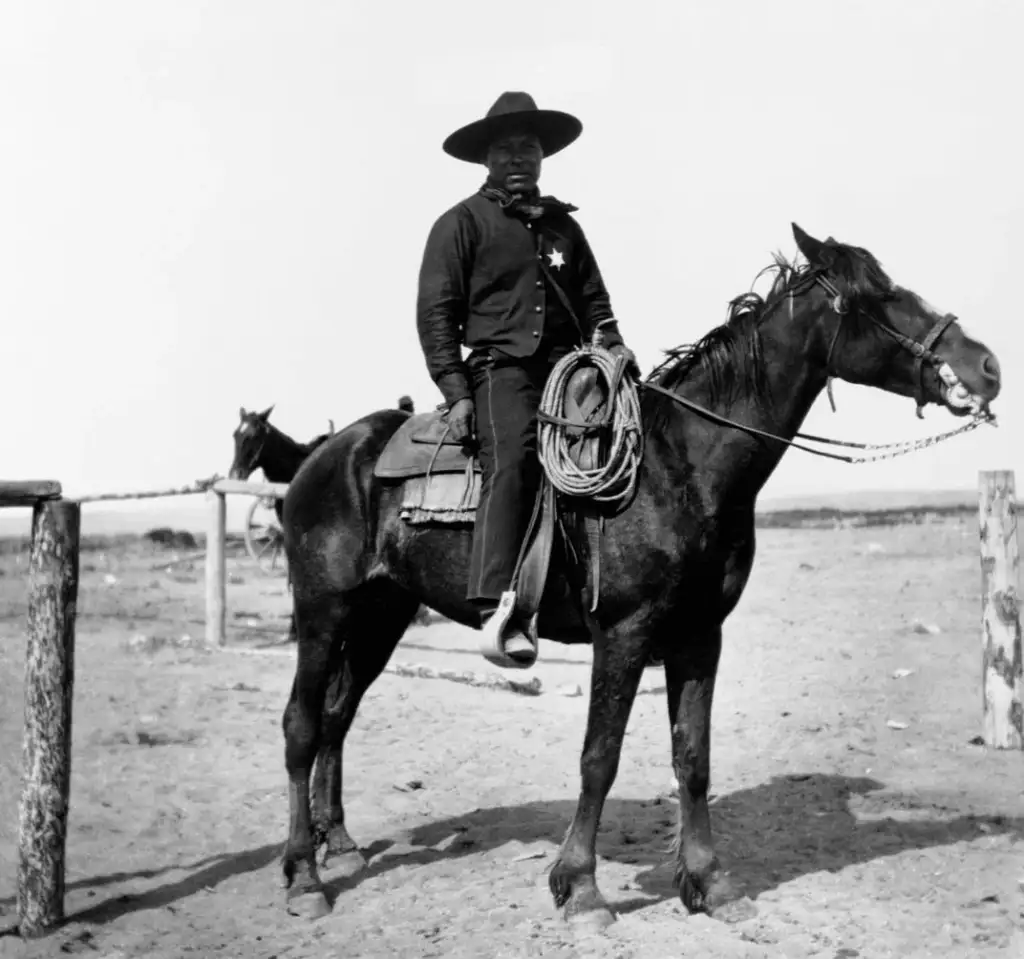 A black-and-white photo of a person dressed as a cowboy, wearing a wide-brimmed hat and a badge, sitting on a dark horse. The scene is set in an open, sandy area with another horse tied to a post in the background.