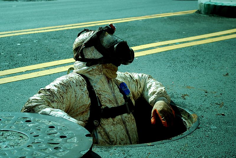 A person in a full protective hazmat suit and gas mask is emerging from a manhole on a paved street. The individual is covered in a messy, muddy substance. The road features double yellow lines.