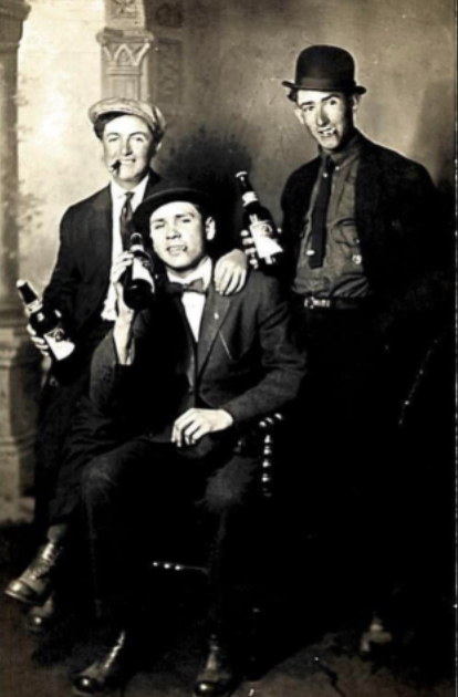 Three men in vintage attire pose with bottles in a sepia-toned photograph. The man on the left sits holding a bottle, while the other two stand, wearing bowler hats and smiling, with one holding a bottle. They appear in a classic, old-fashioned setting.