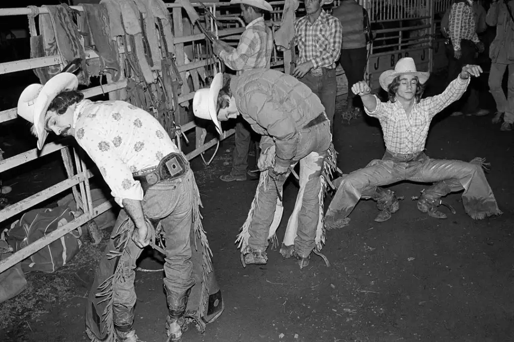 Three cowboys wearing hats and chaps prepare for a rodeo, two standing and adjusting gear while the third is stretching. They are surrounded by rodeo equipment and other people in the dimly lit area.