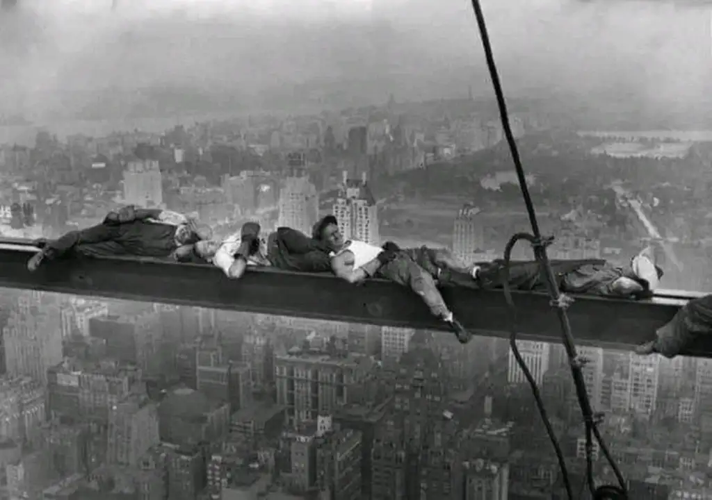 Workers are lying on a steel beam high above a city skyline, surrounded by fog. The image is in black and white, showing the workers resting in a precarious position, embodying historical construction scenes.