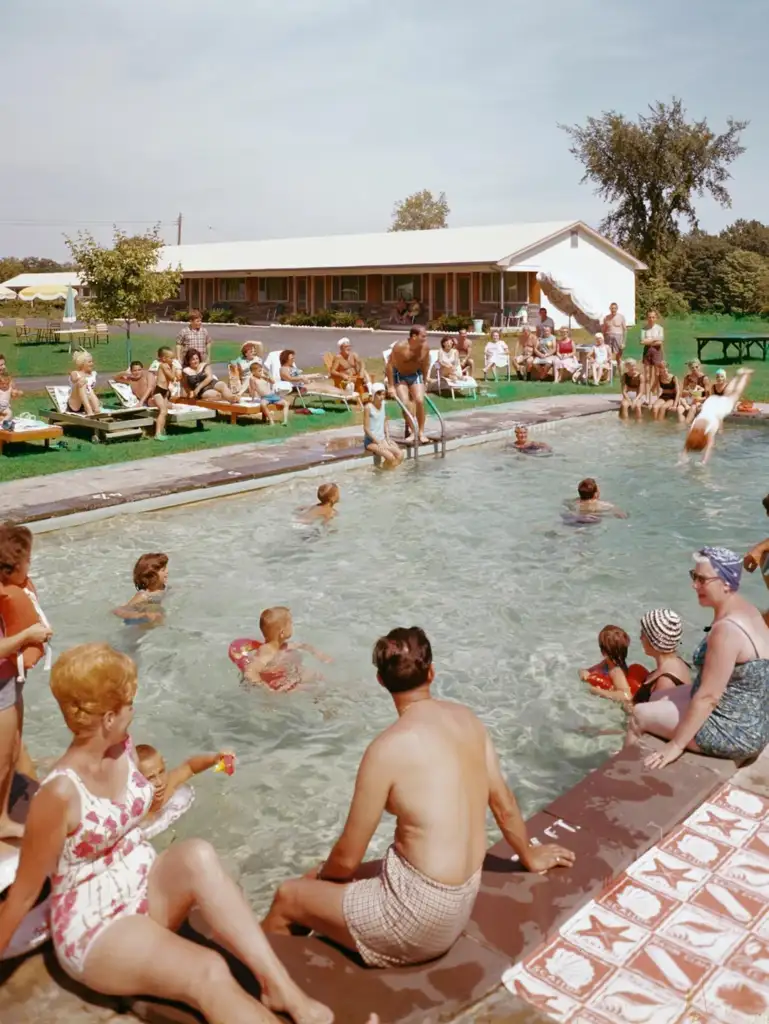 A lively swimming pool scene with adults and children enjoying the water. People are sitting on the edge, some standing in the pool, and more are lounging on chairs nearby. A building in the background adds to the summery, leisure atmosphere.