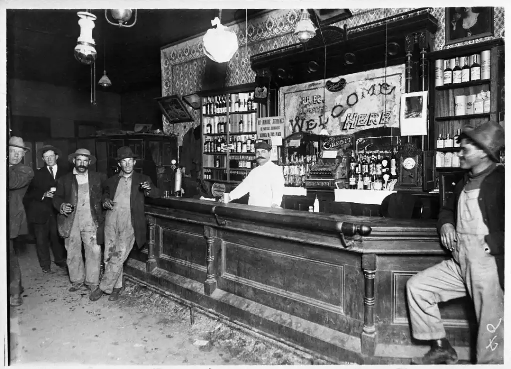 Black and white photo of a historic bar scene. A bartender stands behind a wooden counter with bottles and a "Welcome Here" banner. Four men in hats and work clothes are at the bar, three standing, and one leaning against the counter.