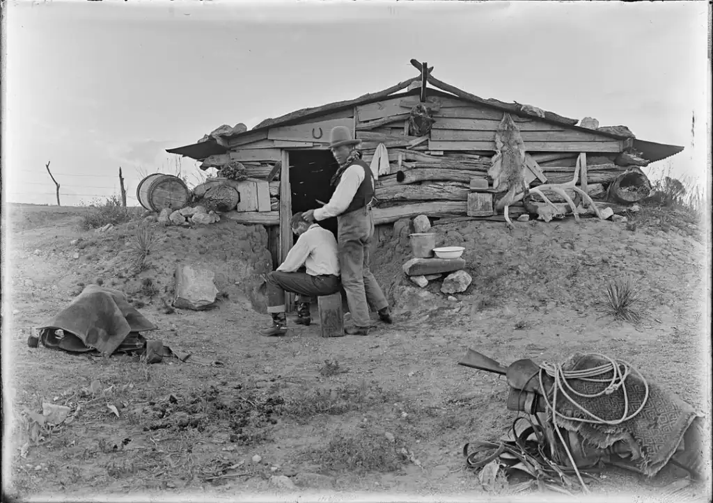 Two men in front of a rustic log cabin in a rural setting. One is seated, hair being cut by the other. Various items like baskets and animal hides adorn the cabin. A saddle and rope lie on the ground nearby. Sparse grass and open sky in the background.