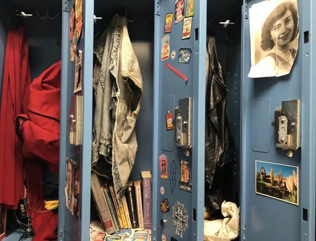 Open lockers with blue doors display personal items. Left locker has a red garment, middle locker has clothing and books, and right locker shows jackets and photos, including a close-up of a smiling woman. Stickers decorate the inside surfaces.