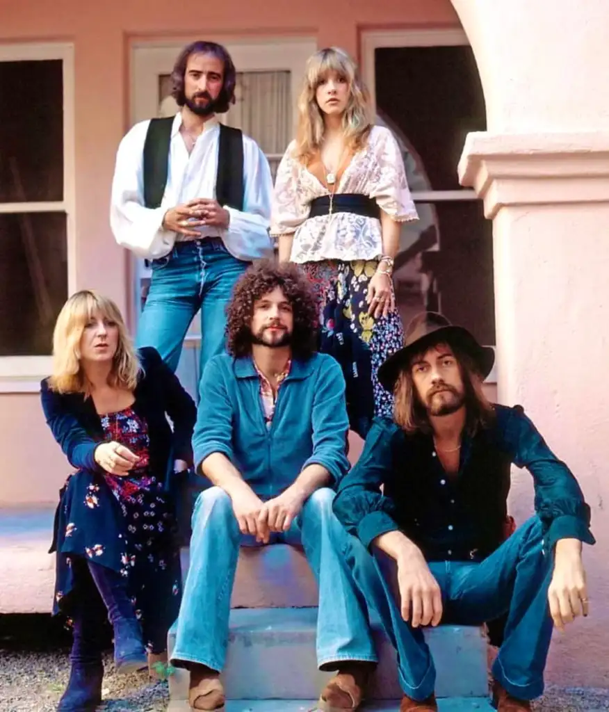 Five people are posed outside a pink building. Two are sitting on steps, three are standing behind. They wear vintage-style clothing, including hats, vests, and floral prints. The mood appears relaxed and stylish.