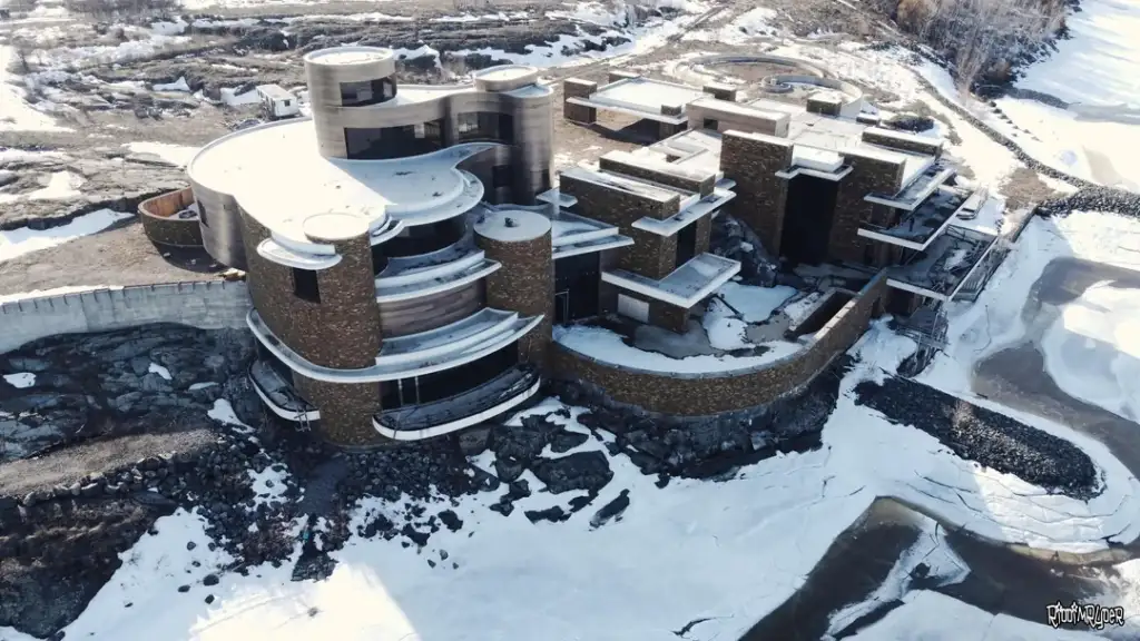 Aerial view of a large, futuristic building with circular and rectangular structures. The building is set on a rocky, snowy landscape, near a partially frozen body of water. The architecture features stone and metal elements.