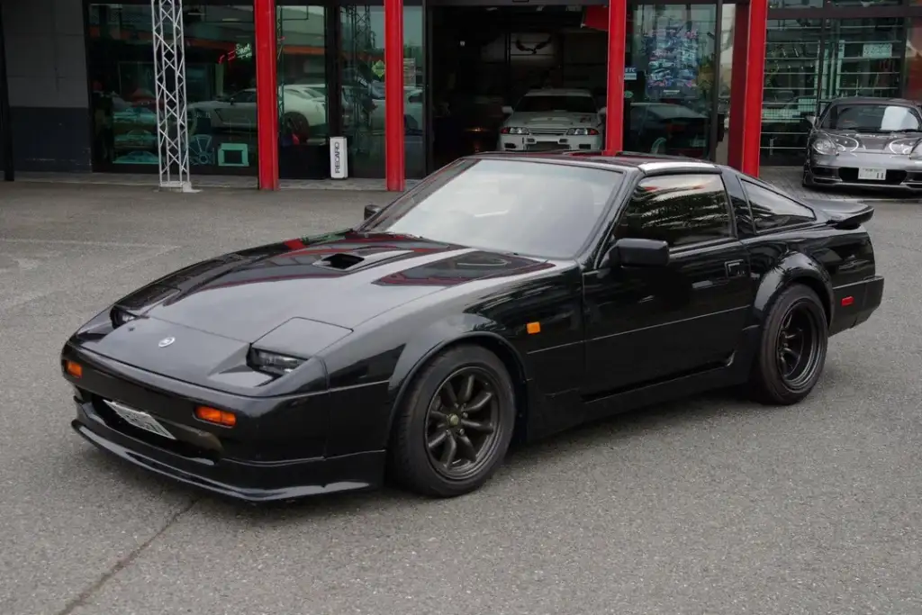 A sleek, black sports car is parked in front of a building. The car has pop-up headlights, a low profile, and dark rims. The building behind has large windows displaying various other vehicles inside.