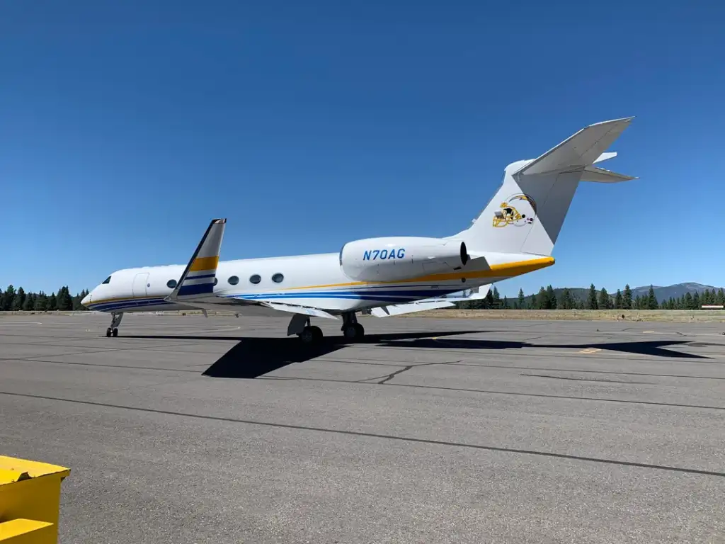 Private jet on a runway under a clear blue sky. The aircraft is white with yellow and blue stripes and has a logo on the tail fin. Mountains are visible in the background, and the runway is surrounded by a few distant trees.