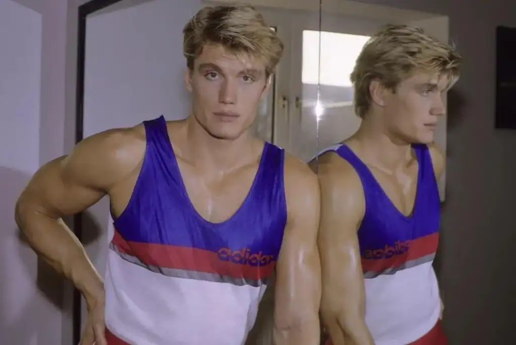 A man with blond hair in a sleeveless Adidas athletic shirt poses confidently indoors. He appears twice in the image, with his reflection visible in a mirror behind him. The shirt features blue, red, and white colors.