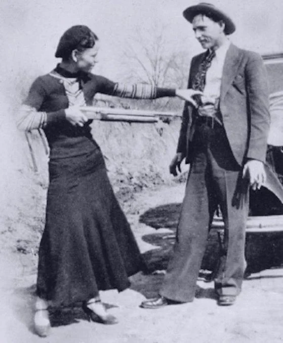 A historical black-and-white photo showing a woman playfully pointing a shotgun at a man. Both are in 1930s attire, standing outdoors near a classic car. The woman wears a long dark dress, and the man is in a suit with a tie and hat.