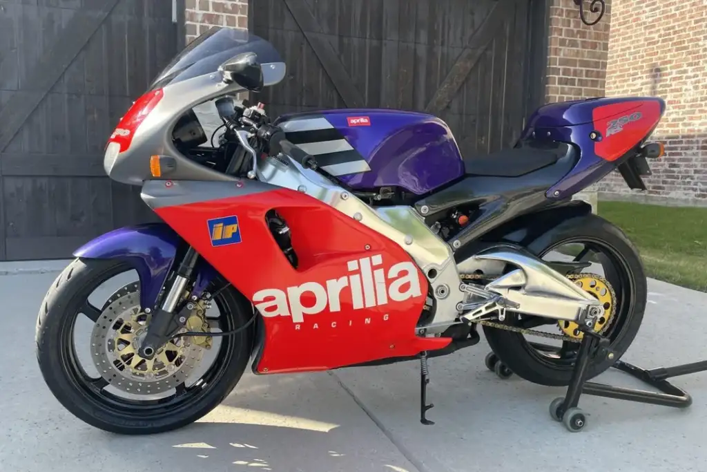 A racing motorcycle with a blue, red, and silver color scheme is parked on a concrete surface. It features "Aprilia Racing" branding on the side, mostly red front section, and visible disc brakes and exhaust system. A brick wall is in the background.