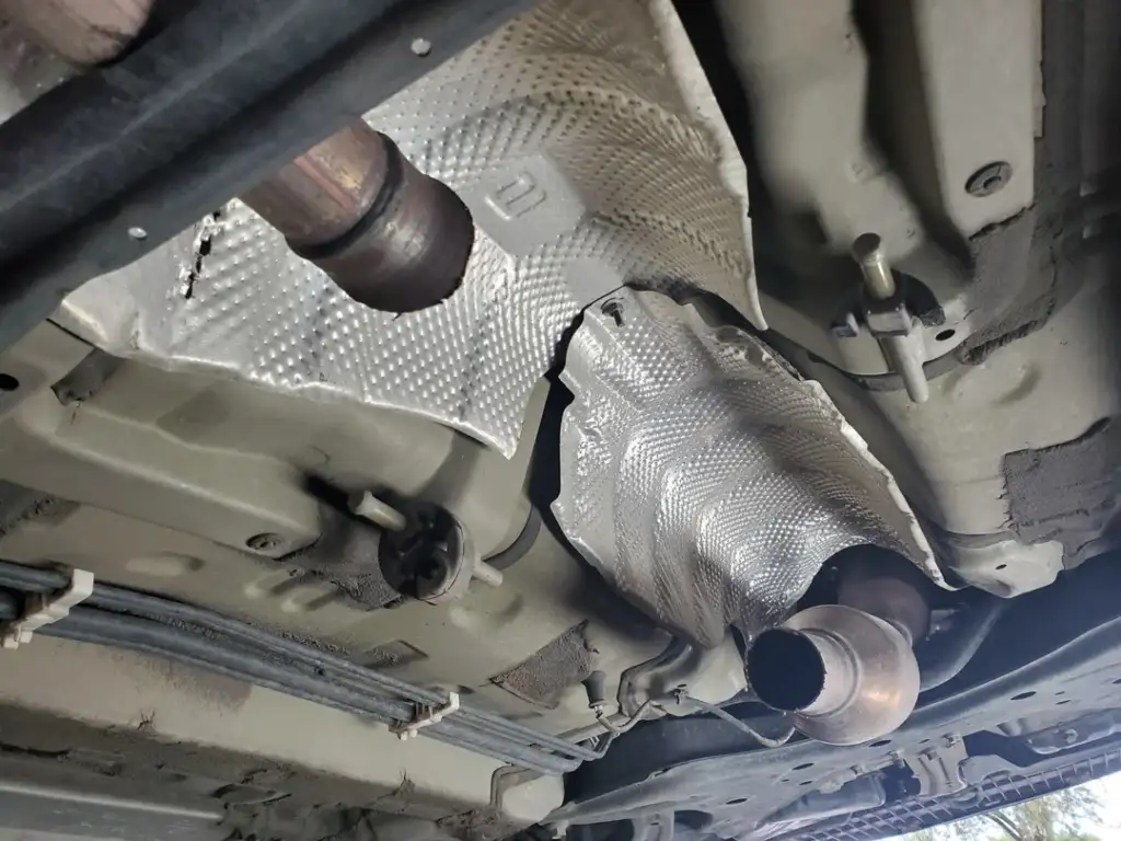 View of a vehicle's underside showing exhaust pipes and a section of metallic protective heat shielding. The surrounding area has visible metal framework and other mechanical components.