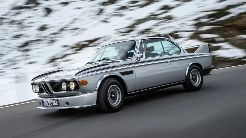 A classic silver BMW car with black detailing is driving along a mountain road. The road is surrounded by snow-covered ground, showcasing a mix of asphalt and snowy terrain. The car is viewed from the side, highlighting its vintage design.