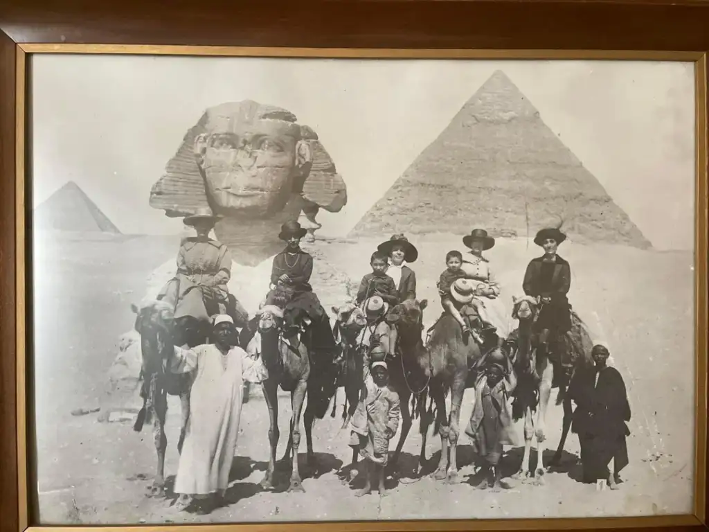 A vintage black-and-white photo shows a group of people posing with camels in front of the Sphinx and pyramids in Egypt. Some are on the camels while others stand beside them, all dressed in early 20th-century attire.