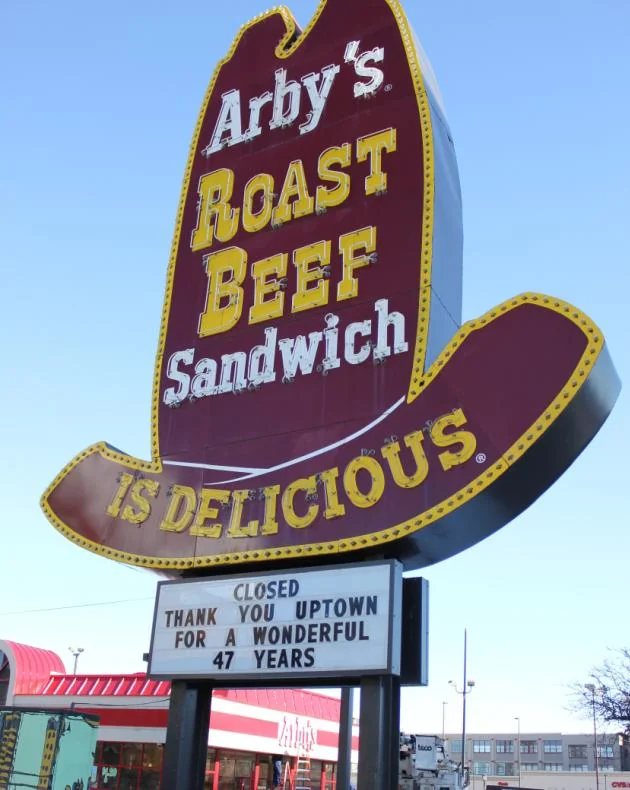 A large Arby's cowboy hat sign reads "Arby's Roast Beef Sandwich is Delicious." Below it, a marquee states, "Closed Thank You Uptown for a Wonderful 47 Years." The sky is clear, and a red-roofed building is visible.