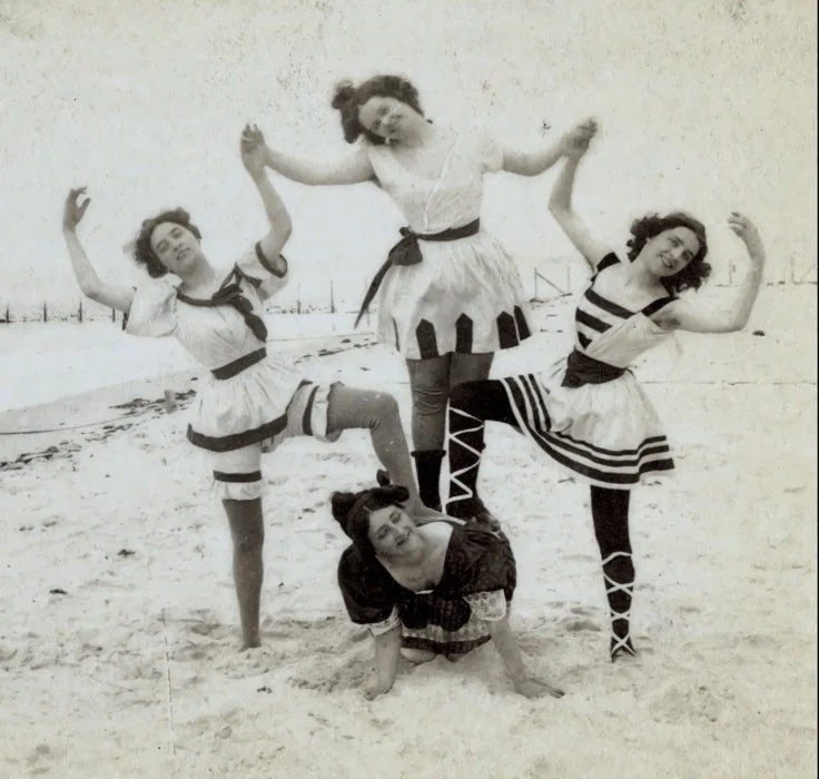 Four women in vintage swimsuits pose playfully on a snowy beach. Three stand with linked arms, while one crouches in front, smiling. The swimwear features stripes and lace-up leggings, contrasting with the wintry background.