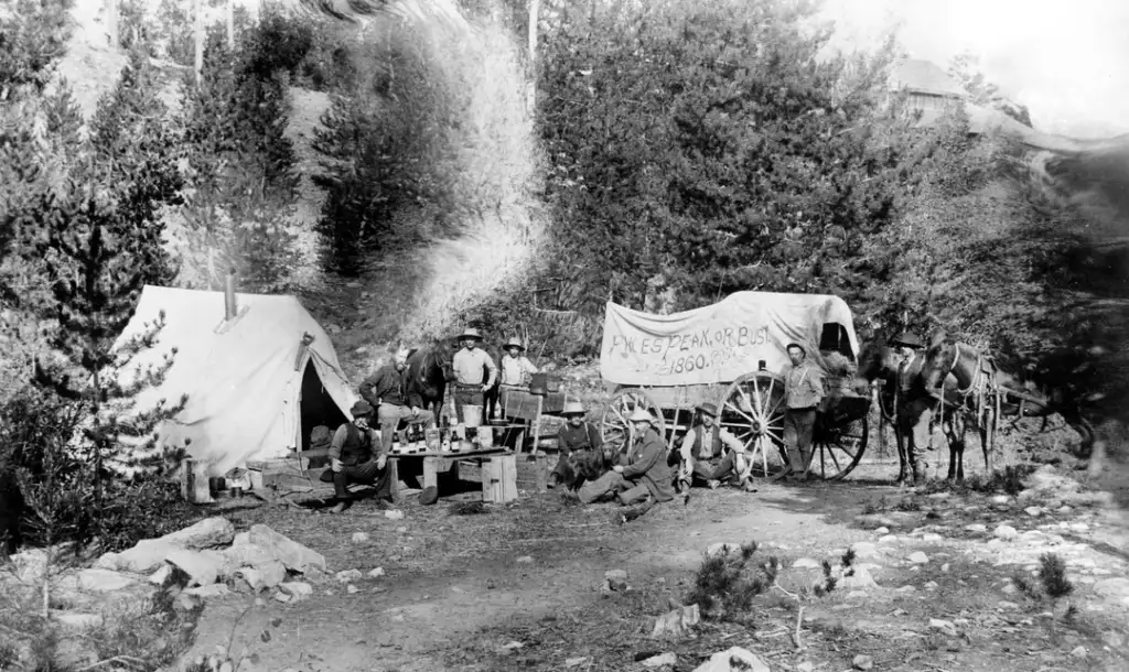 A vintage scene depicts a group of people in a forest camp beside two canvas tents and a covered wagon. A banner reads "Pikes Peak or Bust, 1858." There are several men gathered around camp tables, with horses nearby.