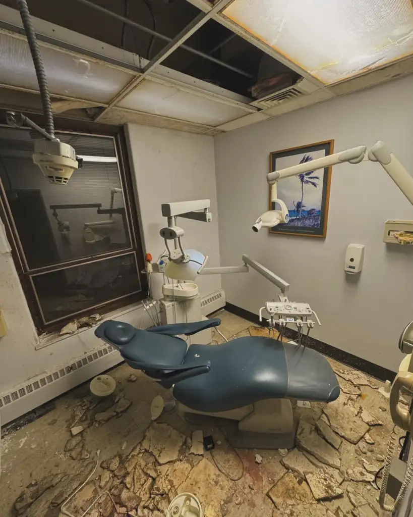 Abandoned dental office with worn-out dental chair and equipment. Ceiling tiles are missing, and debris covers the floor. A painting of a tree hangs on the wall beside a window. The room appears neglected and in disrepair.