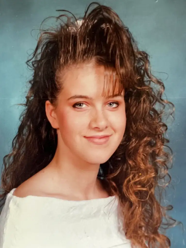 A person with long, curly brown hair styled in a high wave on top, wearing a white off-the-shoulder top, against a blue background, smiling slightly.