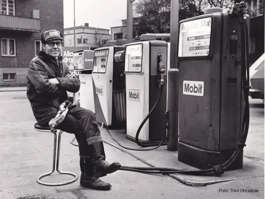 A man wearing a cap and glasses sits on a stool while holding a fuel pump nozzle, smiling. Behind him are three vintage Mobil gas pumps labeled Regular Mix, Premium, and Economy. The scene is set in an outdoor urban area.