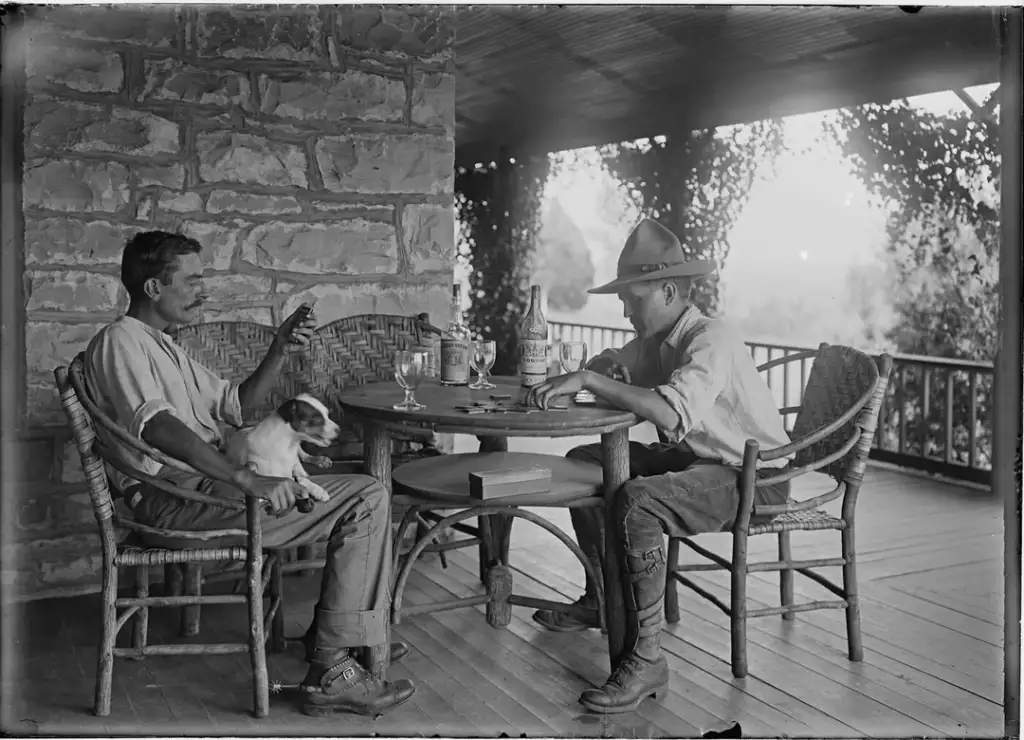 Two men sit at a round table on a covered porch, engaged in conversation. One holds a small dog on his lap, while the other wears a wide-brimmed hat. The porch is lined with wicker chairs and climbing plants. Glasses and a jug complete the setting.