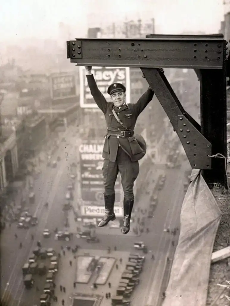 A man in a uniform hangs from a steel beam high above a bustling city street. Below, cars and people are visible, and signs on buildings are partially readable. The scene is depicted in black and white.