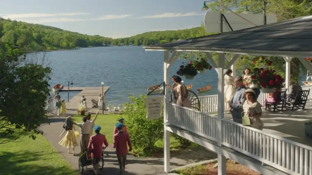 A group of people walk towards a lakeside dock surrounded by lush green trees. Others gather on a covered porch with hanging flowers. A sign reads "Boats Available." The lake and sky are clear on a sunny day.