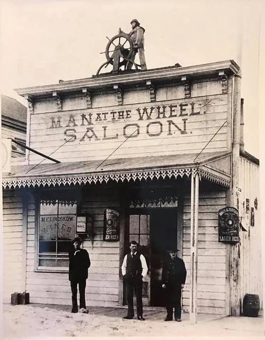 Historical photo of a saloon labeled "Man at the Wheel" with wooden architecture. Three men stand in front of the entrance, and one man is positioned atop the building holding a ship's steering wheel. Signs for ales and lager beer are visible.