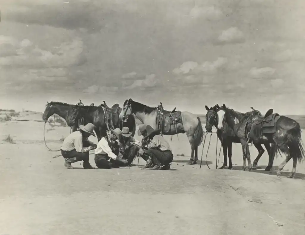 Five men wearing hats crouch on a dirt ground, examining something closely. Behind them, six saddled horses stand in a barren, open landscape under a cloudy sky.