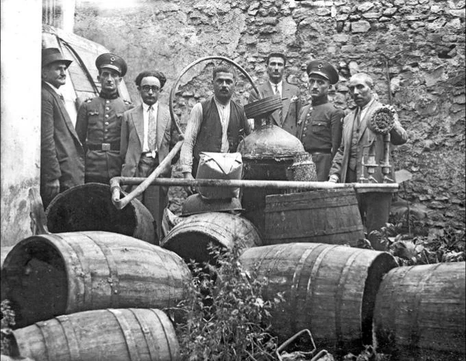 A black and white photo of eight men, some in uniform, standing outside near a stone wall. In front of them are barrels and distillery equipment. The scene appears historical and may relate to a prohibition or enforcement context.