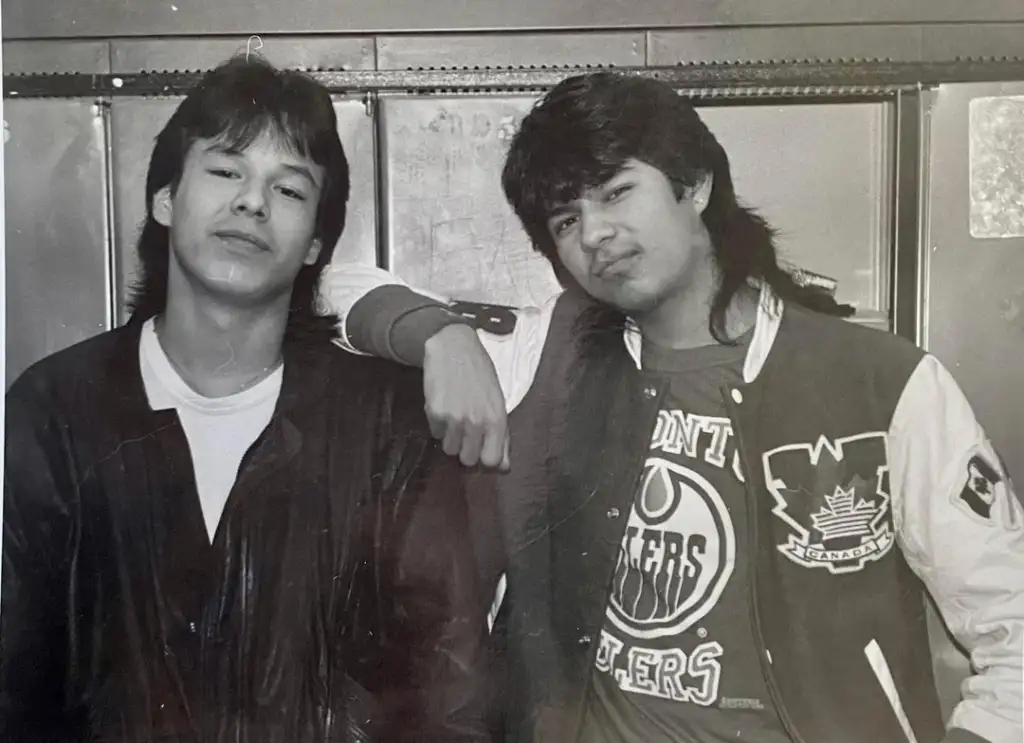 Two young men pose casually, leaning against a surface. One wears a leather jacket, the other a varsity-style jacket with a sports logo. Both have relaxed expressions and wear their hair in a mullet style, typical of 1980s fashion. Black and white image.