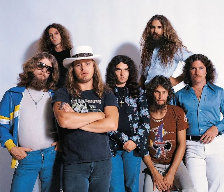 Seven men with long hair stand and pose against a plain white backdrop. One wears a white hat, and others have beards and casual 70s attire including patterned shirts and sunglasses. They look confidently toward the camera.