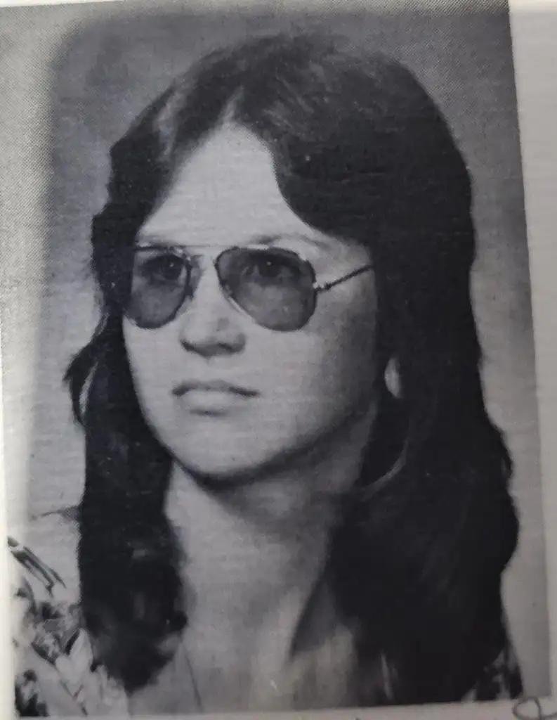 A black and white portrait of a young person with long hair, wearing aviator sunglasses. They are facing slightly to the left with a neutral expression, and are dressed in a patterned shirt.