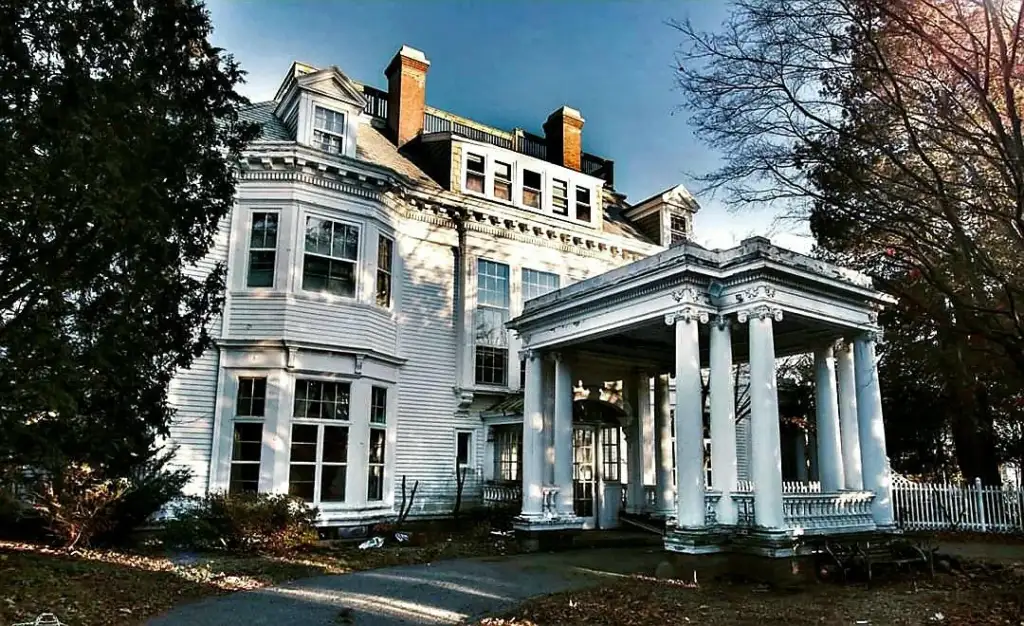 A large, historic mansion with white wooden siding, tall columns, and multiple windows stands under a blue sky. The building features a portico and mature trees are visible around the property.