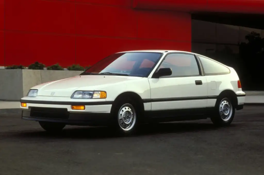 A white hatchback car, likely from the late 1980s or early 1990s, is parked on a street. It has a compact design with black trim and visible headlights. The background features a red wall and some greenery.