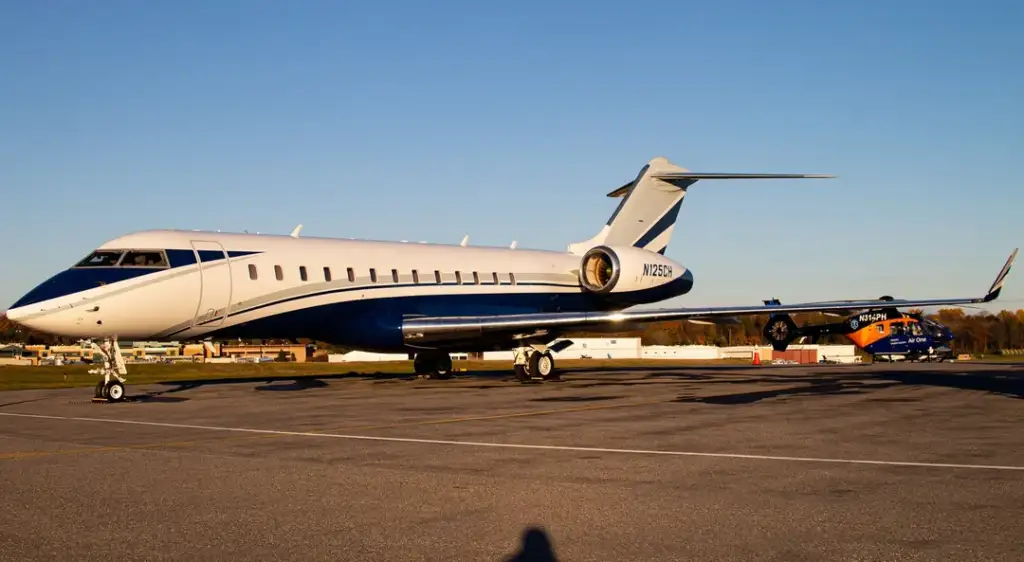 A sleek private jet with a blue and white color scheme is parked on an airstrip under a clear blue sky. In the background, a helicopter is visible on the tarmac. The sun casts long shadows in the foreground.