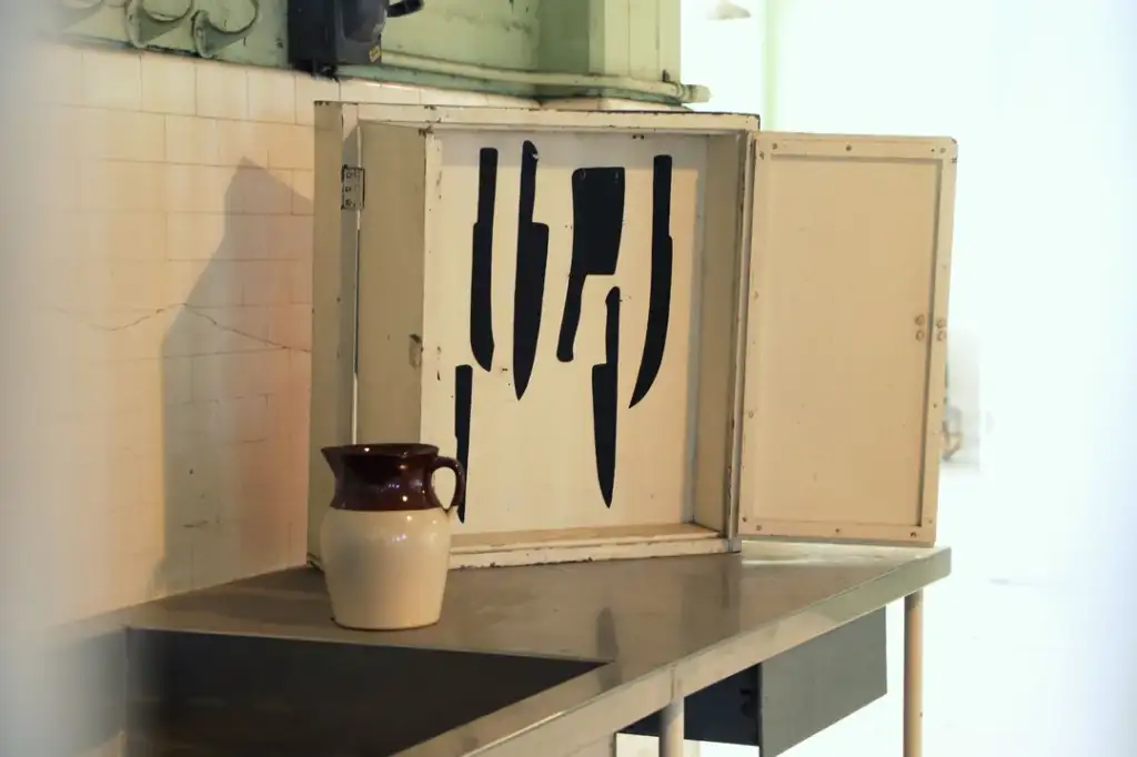 A kitchen scene featuring an open metal cabinet on a table, displaying several large knives hanging inside. A ceramic pitcher with a brown top and white bottom sits nearby. The background is softly lit.