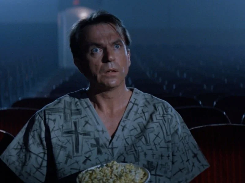 A man with wide eyes, wearing a patterned shirt, sits in an empty theater holding a bowl of popcorn. The dimly lit setting creates a suspenseful atmosphere.