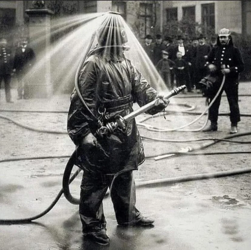 A firefighter in an early 20th-century protective suit stands holding a hose, with water spraying over their helmet. Onlookers, including fellow firefighters, watch the demonstration. Hoses lie on the ground in a city setting.