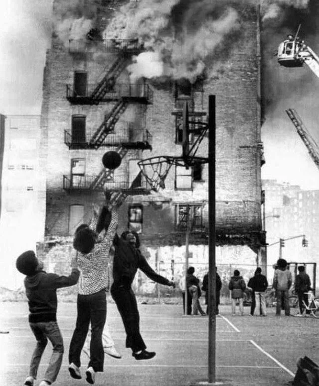 A black and white photo of children playing basketball on an outdoor court. Behind them, a building is engulfed in flames, with smoke billowing out. A firetruck ladder is extended toward the building as onlookers watch.