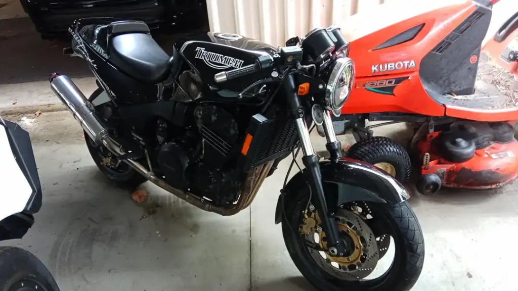 A black Triumph motorcycle is parked in a garage, featuring a prominent exhaust pipe and circular headlight. Behind it is an orange Kubota riding lawn mower, partially visible. The floor is concrete, and other equipment surrounds the motorcycle.