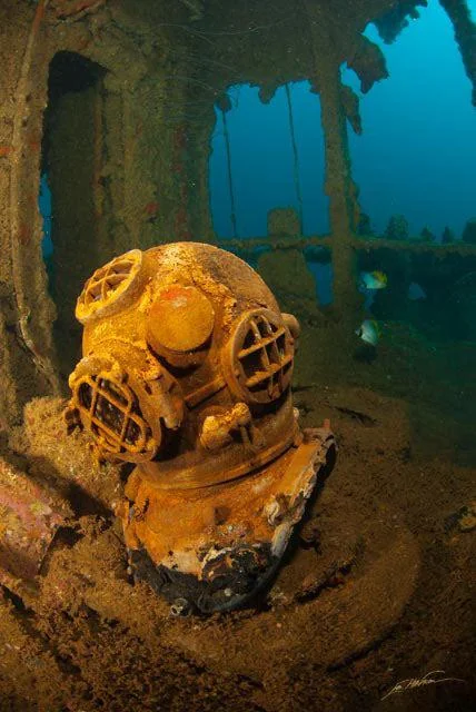 An old, rusted diving helmet sits on a sandy seabed inside a sunken shipwreck. The metal is corroded, with round, grated viewports. The surrounding water is a deep blue, with fish swimming in the background.