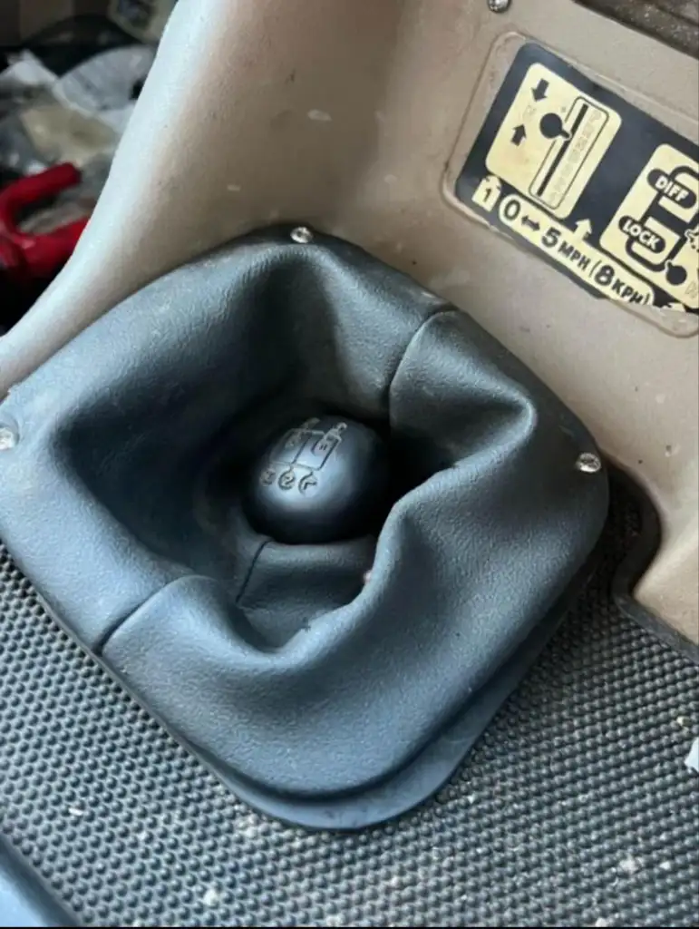 Close-up of a vehicle's gear shifter covered with a soft black leather boot. The gear pattern is visible on the round shift knob. A metal plate nearby shows a diagram related to differential lock instructions. The floor is covered in textured material.