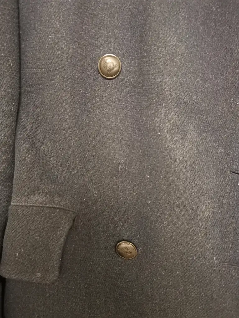 Close-up of a dark gray woolen coat with two metallic buttons. The fabric has a slightly textured appearance, and a part of a pocket flap is visible at the bottom left.