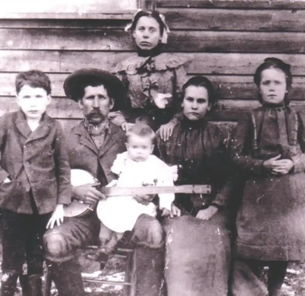 A vintage black and white photo of a family posing in front of a wooden wall. Six people are seen: a man with a hat holding a banjo, a woman holding a baby, and three children standing around them. The attire suggests an early 20th-century setting.