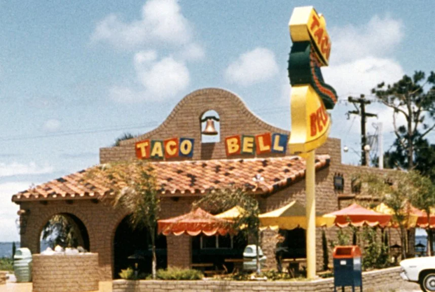 Vintage Taco Bell building with rustic architecture, featuring a tan brick facade, arched entryway, and a colorful sign. Bright umbrellas adorn the patio area, and a large decorative bell is mounted on the roof. Blue sky and trees in the background.