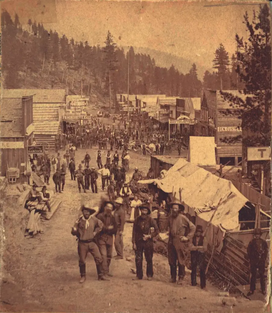 A sepia-toned photograph of an old west town with people walking along a dusty street. Wooden buildings line the sides, and a forested hill is visible in the background. Several figures wear hats and some appear to be engaged in conversation.