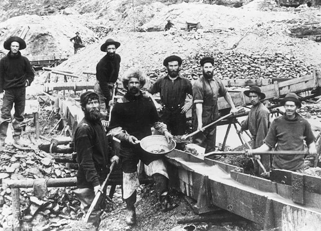 Black and white photo of eight gold prospectors during the Klondike Gold Rush. They stand on rocky terrain with tools like pans and shovels. A wooden sluice box is visible, and the men wear period-appropriate rugged clothing and hats.